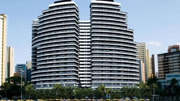 Un hotel moderno de gran altura con fachada curva, situado cerca de una playa con sombrillas y palmeras.