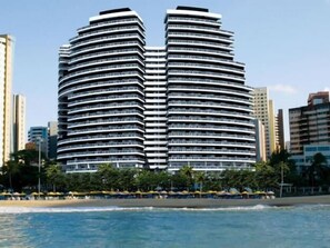 A high-rise hotel with a curved facade, located near a beach with umbrellas and palm trees.
