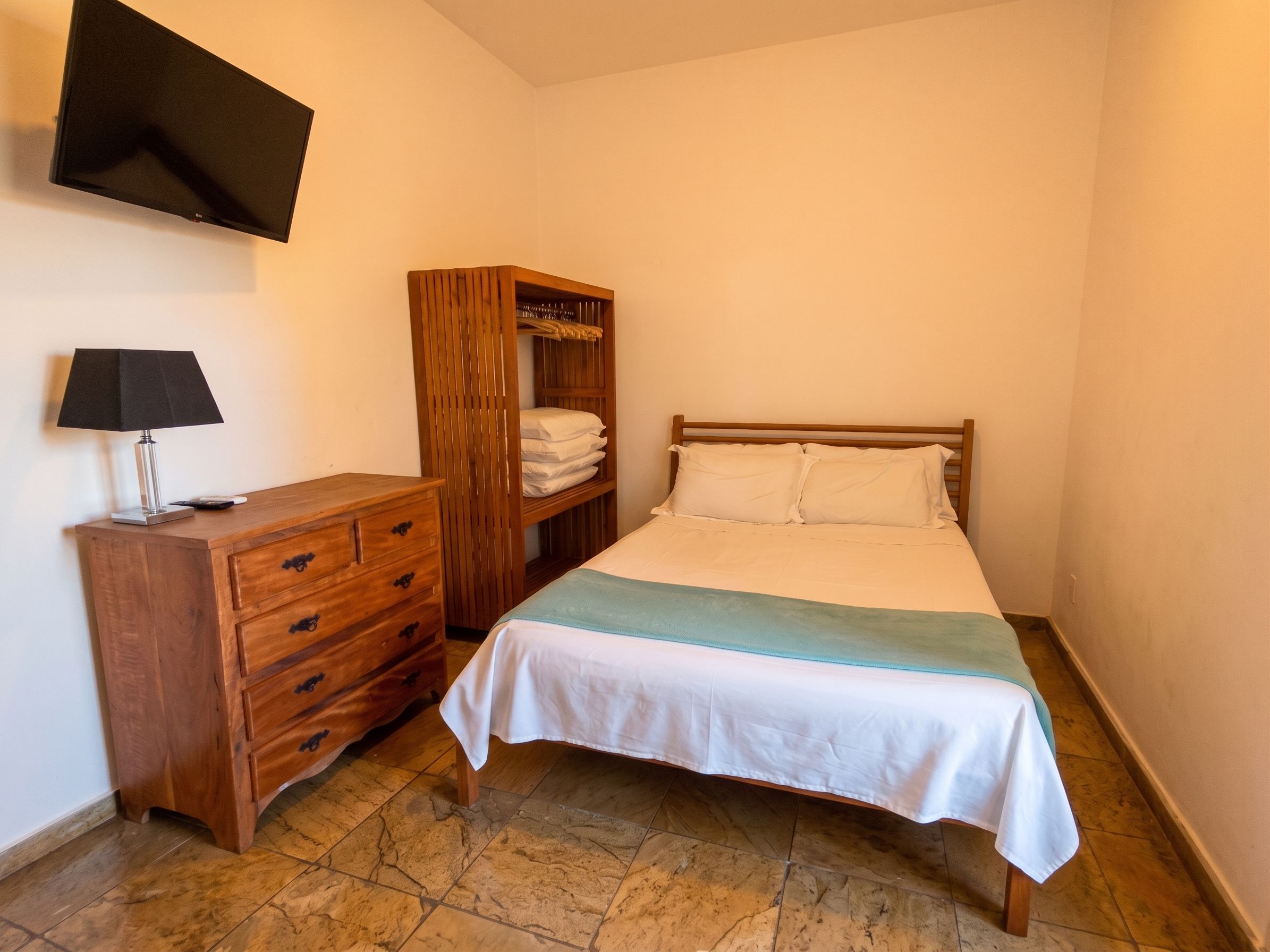A bedroom with a bed, a wooden dresser, a wardrobe, and a flat-screen TV mounted on the wall.