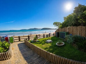 Vista de la playa con una cerca de madera, un área con césped y plantas, y un cielo despejado.