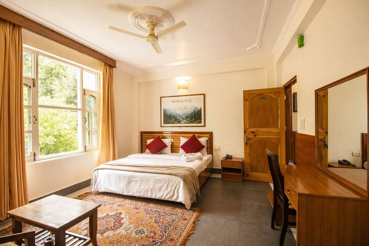 Una camera d'albergo con un letto, una scrivania, una sedia, una lampada da scrivania, un telefono, uno specchio, una ventola a soffitto, una finestra con tende e un quadro appeso al muro.