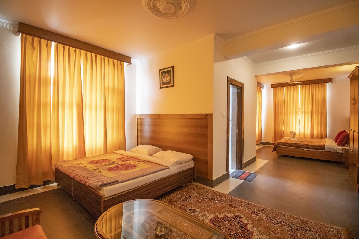Una camera d'hotel con un letto di legno, un tavolino e una sedia. Si vede una porta che dà su un'altra stanza.