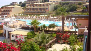 A resort with a swimming pool surrounded by lounge chairs and umbrellas, a palm tree, and a view of hotel buildings in the background. - Luxurious Villa in Apostolata 5★ resort, 3BR, sea views, free parking! (Skala)