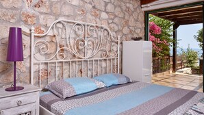 A bedroom with a stone wall, a metal bed frame, a nightstand with a lamp, and a view of a garden through an open door. - Luxurious Villa in Apostolata 5★ resort, 3BR, sea views, free parking! (Skala)