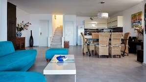 A modern living room with a sofa, a coffee table, and a dining area with wicker chairs. A staircase leads to the kitchen in the background. - Luxurious Villa in Apostolata 5★ resort, 3BR, sea views, free parking! (Skala)
