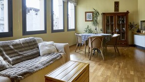 A living room with a sofa, wooden coffee table, and a dining table with chairs. There are large windows with curtains, a cabinet with glass doors, and a plant. - Amazing duplex near the cathedral in the heart to Girona (Gerona)