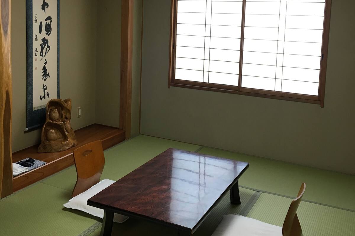 A traditional Japanese room with tatami flooring, a low table, a wooden chair, a calligraphy scroll, and a shoji screen window.