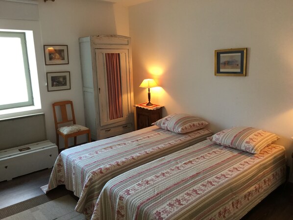 A bedroom with two beds, a chair, a wardrobe, a lamp, and framed pictures on the wall.