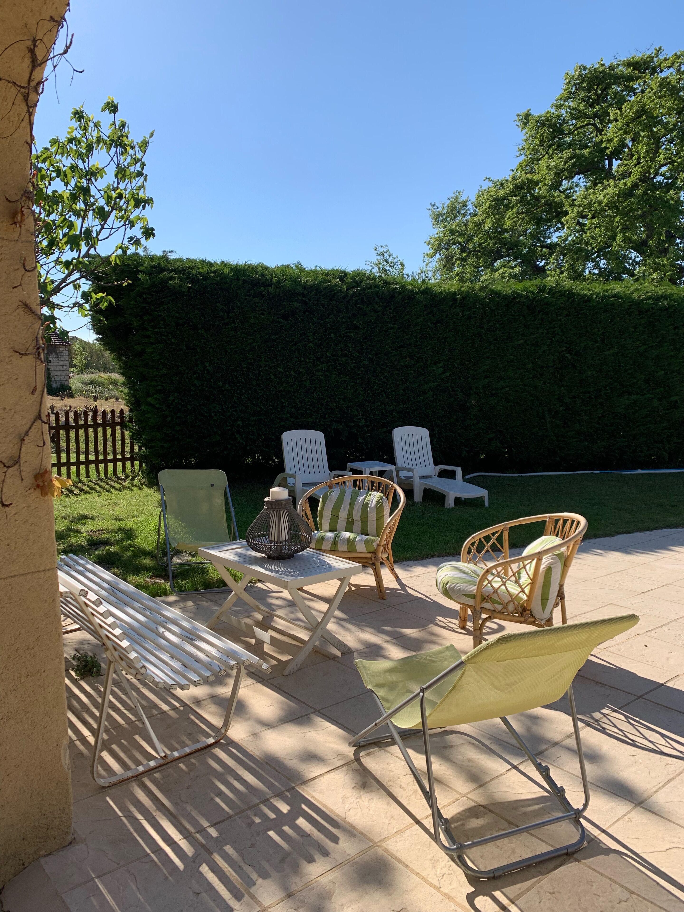 Un patio al aire libre con sillas de mimbre, una mesa con un florero, y un seto bien cuidado.