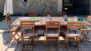 Un espacio para comer al aire libre con un muro de piedra de fondo, una mesa y sillas de madera, y una ventana pequeña.