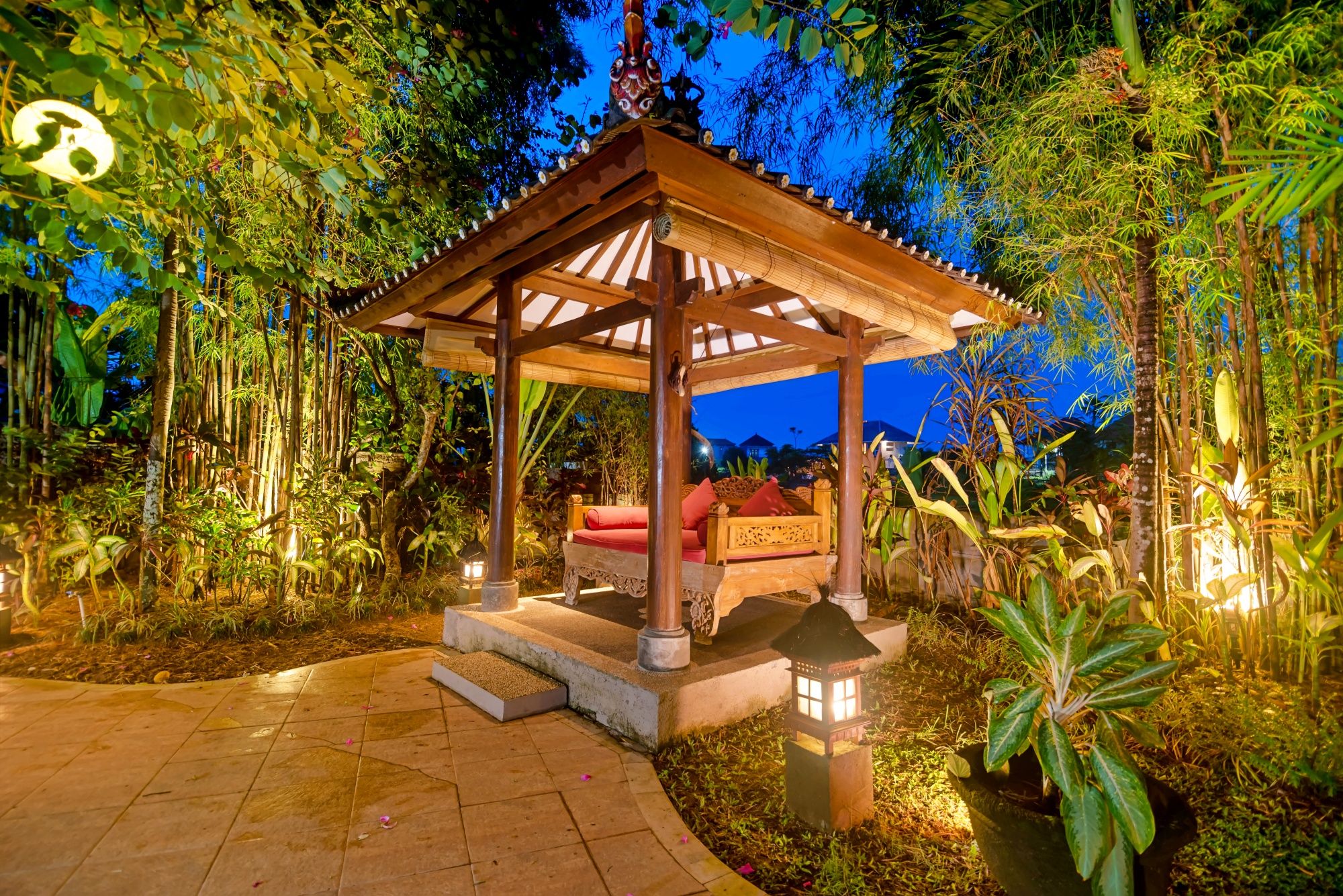 A wooden gazebo with a bench, surrounded by lush greenery and a paved walkway.