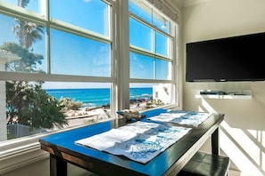 A room with a large window overlooking the ocean, a flat-screen TV mounted on the wall, and a table with a blue tablecloth.