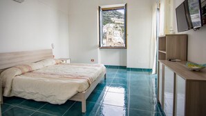 A bedroom with a bed, a desk, a TV, and a window with a view of buildings.