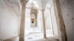 A narrow, arched corridor with stone columns and a tiled floor, leading to a door with a window.