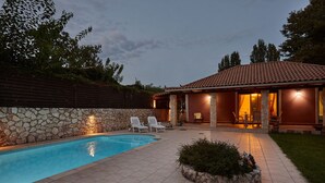 Una piscina amb una paret de pedra, un porxo cobert amb cadires i un vestíbul ben il·luminat.