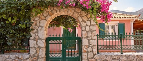 A stone archway with a green gate, leading to a building with a pink facade and green shutters.