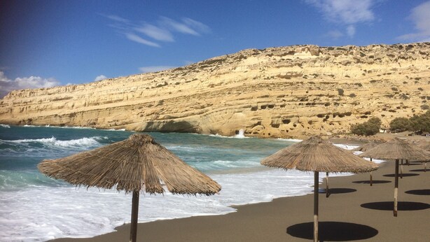 Une plage avec des parasols en chaume et une falaise rocheuse en arrière-plan.