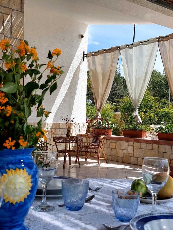 An outdoor patio with a table set for dining, featuring a blue vase with flowers, a glass wine bottle, and a fruit bowl.