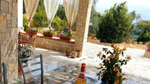 Un área de comedor al aire libre con una mesa dispuesta para dos personas, destacando una botella de vino, un florero con flores y un cuenco de pan y tomates.