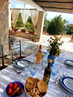An outdoor dining area with a table set for two, featuring a bottle of wine, a vase with flowers, and a bowl of bread and tomatoes.