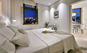 A modern bedroom with a large bed, a view of a cityscape at night, a dining area with a table and chairs, and a small plant on a side table. - Thission Luxury Homes - Acropolis Residence by K&K (Athina)