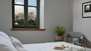 Ein Schlafzimmer mit Blick auf die Akropolis in Athen, ein Bett, ein Sessel, eine Pflanze und ein gerahmtes Bild an der Wand.