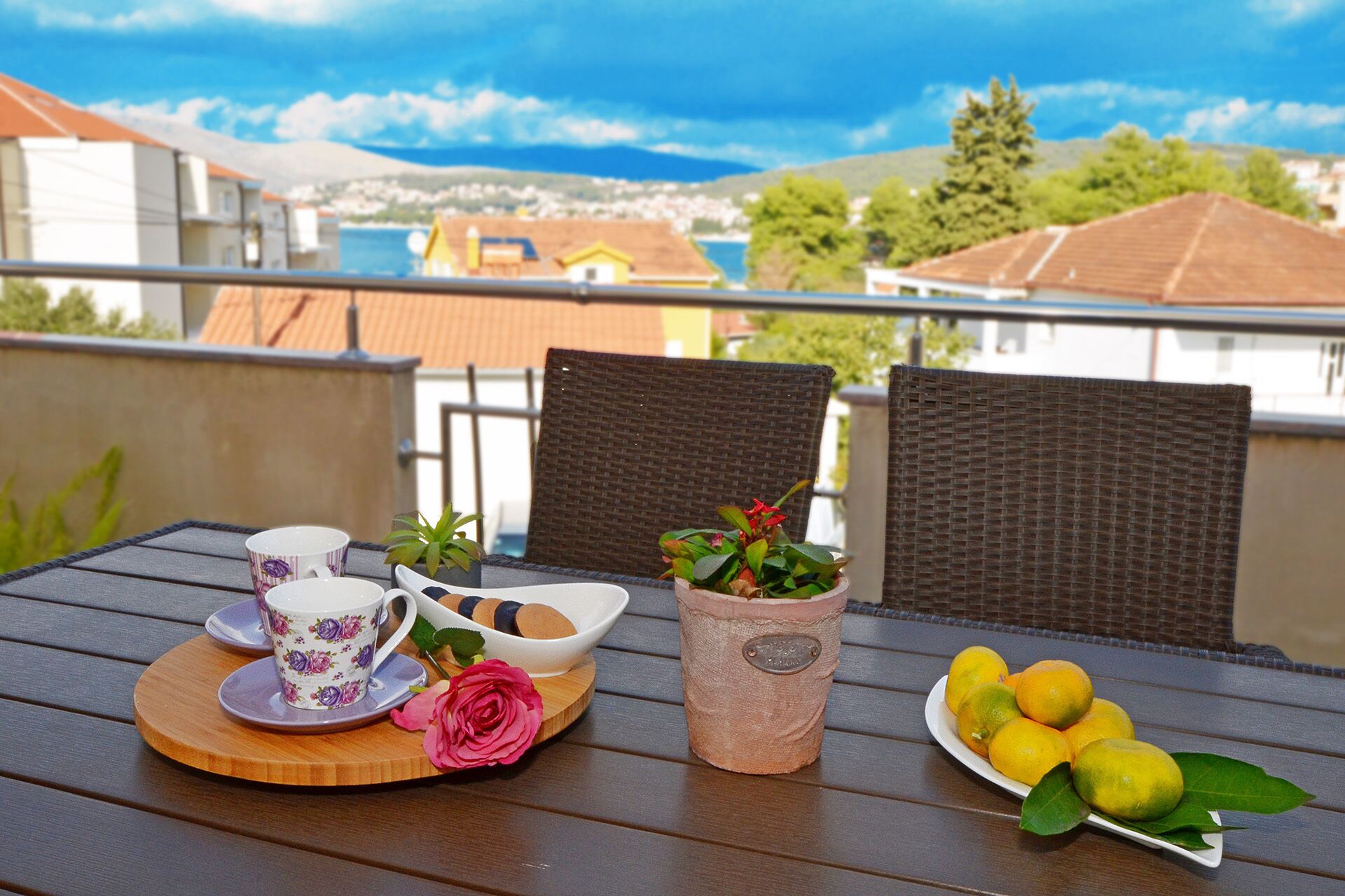A balcony with a table set for tea, featuring a floral teacup, a rose, and a potted plant.