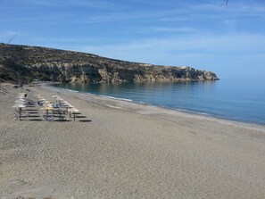 Ein Strand mit Reihen von Sonnenschirmen und Liegestühlen, ein ruhiges Meer und felsige Klippen im Hintergrund.
