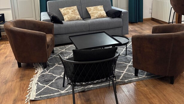 Ein Wohnzimmer mit grauem Sofa, zwei braunen Sesseln, einem schwarzen Couchtisch und einem gemusterten Teppich.
