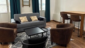 Ein Wohnzimmer mit grauem Sofa, zwei braunen Sesseln, einem schwarzen Couchtisch, einem Holzesstisch mit vier Stühlen und einem großen Fenster mit Vorhängen.