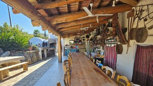 Un espacio exterior para comer con una mesa de madera larga y sillas, piso de baldosas de piedra y techo de madera rústica.