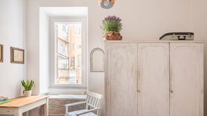 Quarto com escrivaninha branca, cadeira branca, janela com vista para edifícios, guarda-roupa branco e planta em vaso.