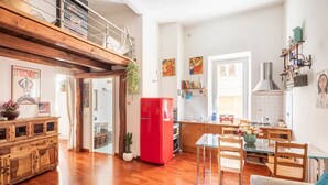 Uma sala de estar ampla com piso de madeira, uma geladeira vermelha, uma mesa de jantar de vidro e um sofá.