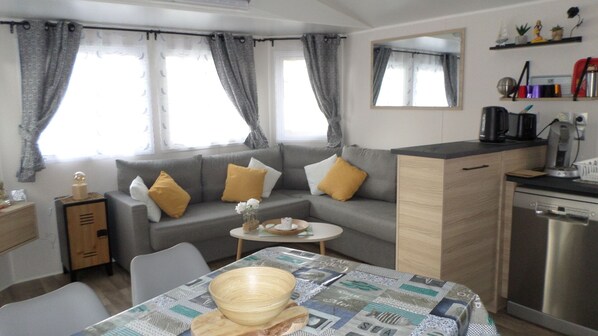 Un espacio habitable compacto con sofá, una pequeña mesa de comedor, una zona de cocina con lavavajillas y un espejo sobre el mostrador.