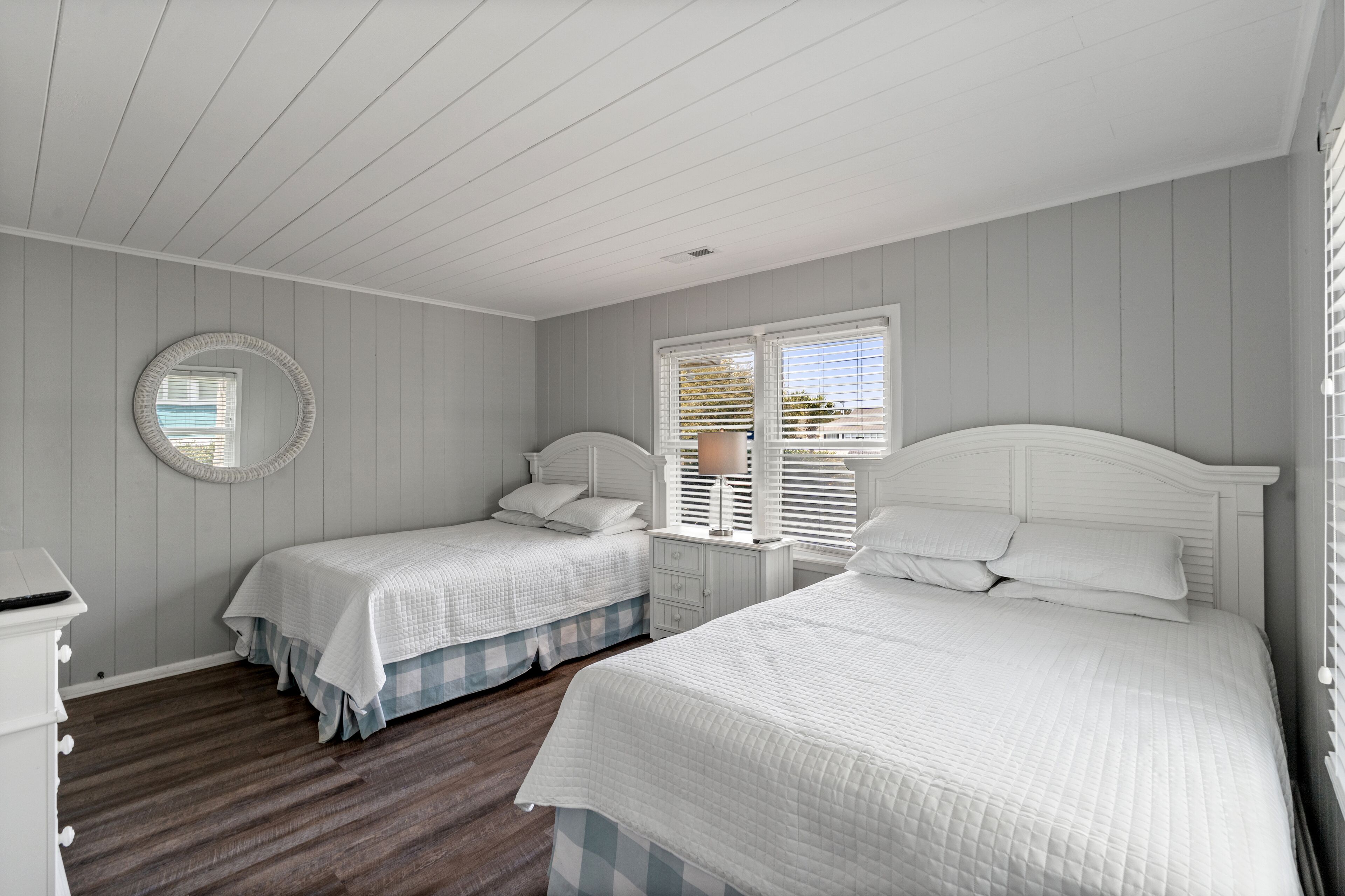 A bedroom with two single beds, a dresser, a round mirror, and a window with blinds.