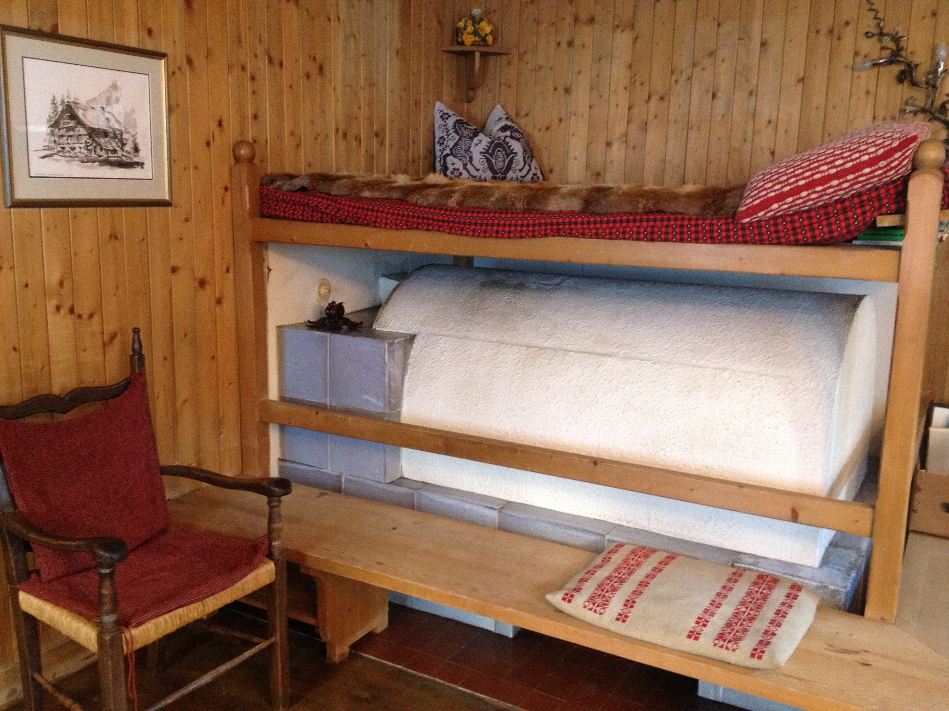 Una cama litera con colcha a cuadros rojo y negro, una silla de madera con cojín rojo, y un cuadro en la pared.