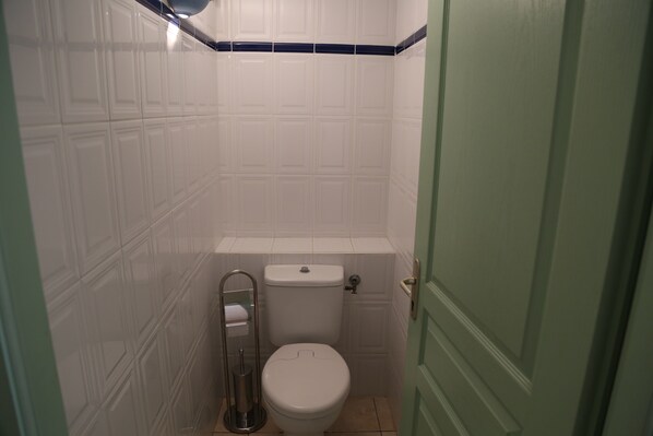 Un baño pequeño con azulejos blancos, un inodoro, un pasamanos metálico y una puerta verde.