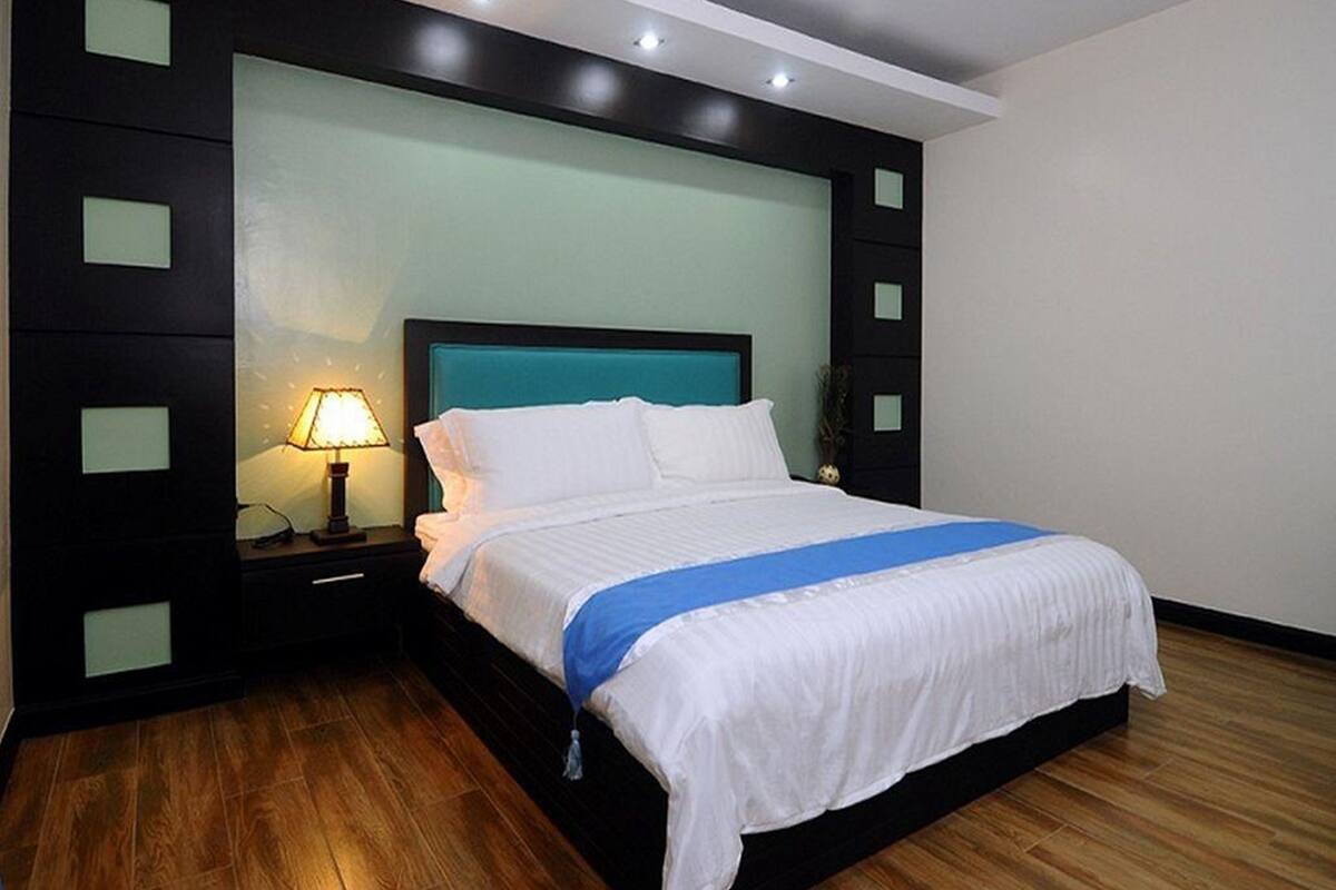 A modern bedroom with a large bed, a nightstand with a lamp, and a dark headboard with light panels.