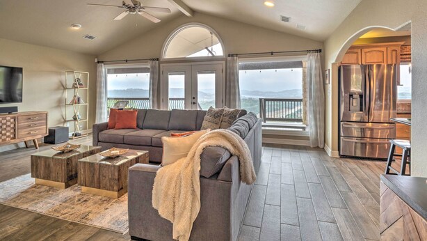 A spacious living room with a gray sofa, wooden coffee tables, and a large window offering a view of the mountains. - Deck & Lake Travis Views: Hill Country Hideaway! (Jonestown)