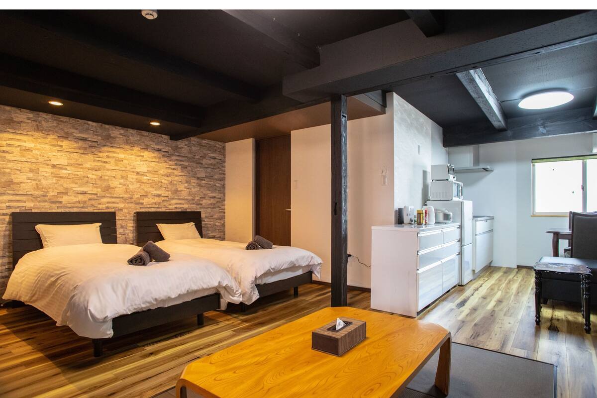 A hotel room with two beds, a wooden table, a kitchenette, and a stone wall.