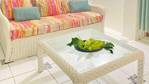 A living room with a white wicker sofa, colorful cushions, a glass coffee table with green plants, and two floral paintings on the wall. - Cozy Apartments close to beach, bars and restaurants (Rodney Bay)