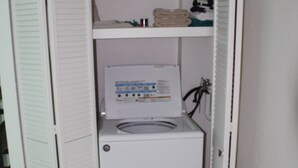 A compact, white laundry room with a front-loading washing machine and a shelf above it. - Cozy Apartments close to beach, bars and restaurants (Rodney Bay)