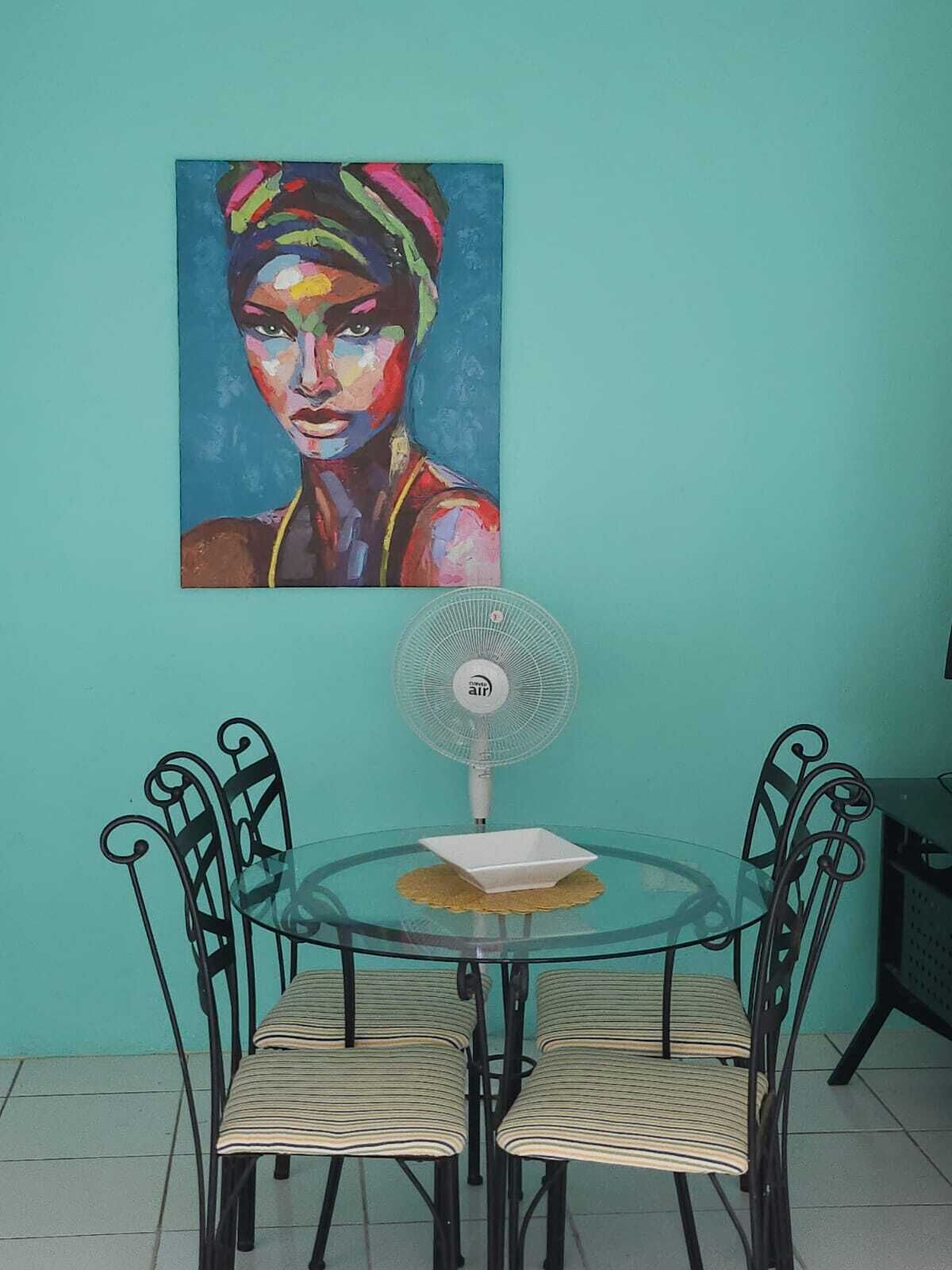 A dining area with a glass table surrounded by four chairs, a white fan, and a colorful painting on the wall.