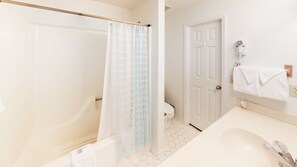A bathroom with a bathtub, a shower curtain, a toilet, a sink, and a towel hanging on the wall. - Golf Villa | Sunset Beach | Live the Golf Life! (CV2B) (Sunset Beach)