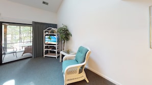 A hotel room with a large window, a wicker chair, a small table, and a TV on a white stand. - Golf Villa | Sunset Beach | Live the Golf Life! (CV2B) (Sunset Beach)