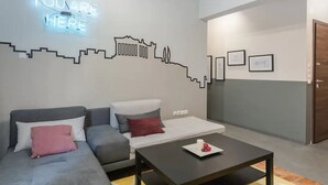 A modern living room with a grey sofa, a small table with a plate of strawberries, a neon sign on the wall, and a cityscape mural.