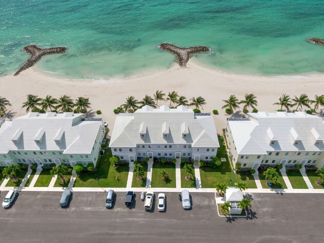 A beachfront hotel with three-story buildings, a parking lot, and a sandy beach with palm trees.