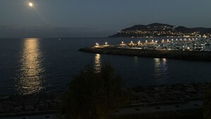 A coastal marina at night with boats docked, a stone breakwater, and a distant illuminated town.