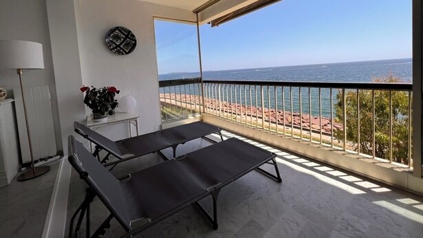 A balcony with two sun loungers, a small table with a vase of flowers, and a view of the sea and beach.