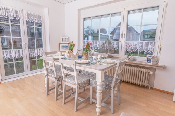 A dining area with a table set for four, surrounded by chairs, large windows with white curtains, and a view of the outside.